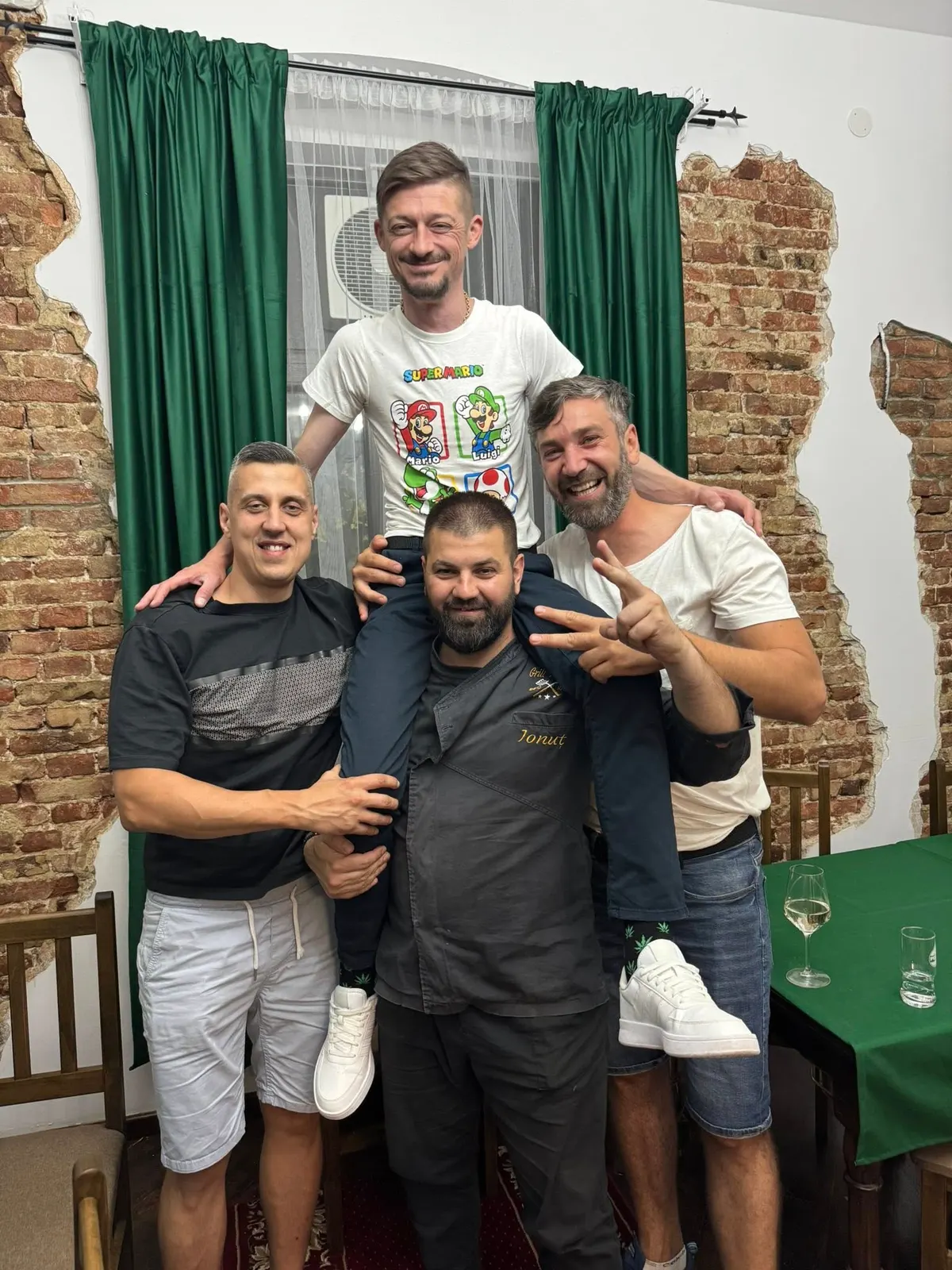 Ion Potîrniche și trei prieteni în salonul restaurantului, în fața peretelui de cărămidă, unul ridicat pe umerii celorlalți, toți râzând