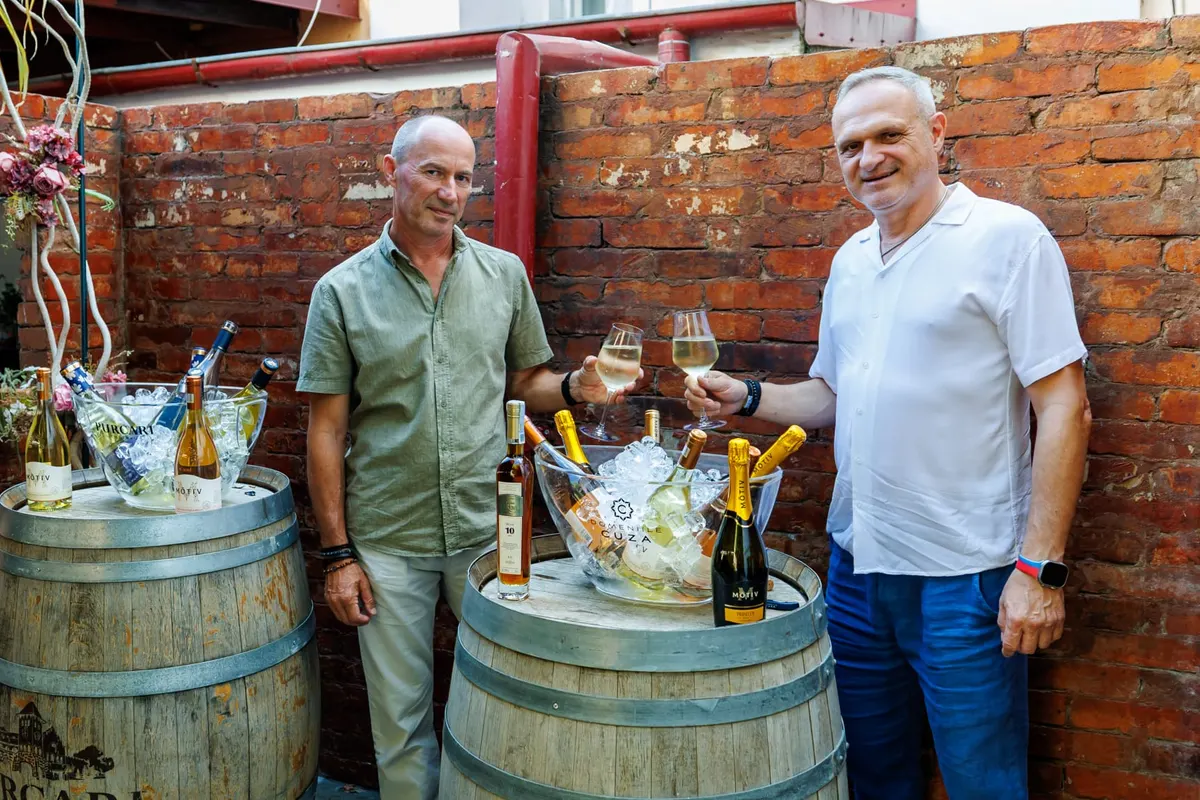 Two men holding glasses of white wine in front of barrels with bottles and ice buckets, against a brick wall