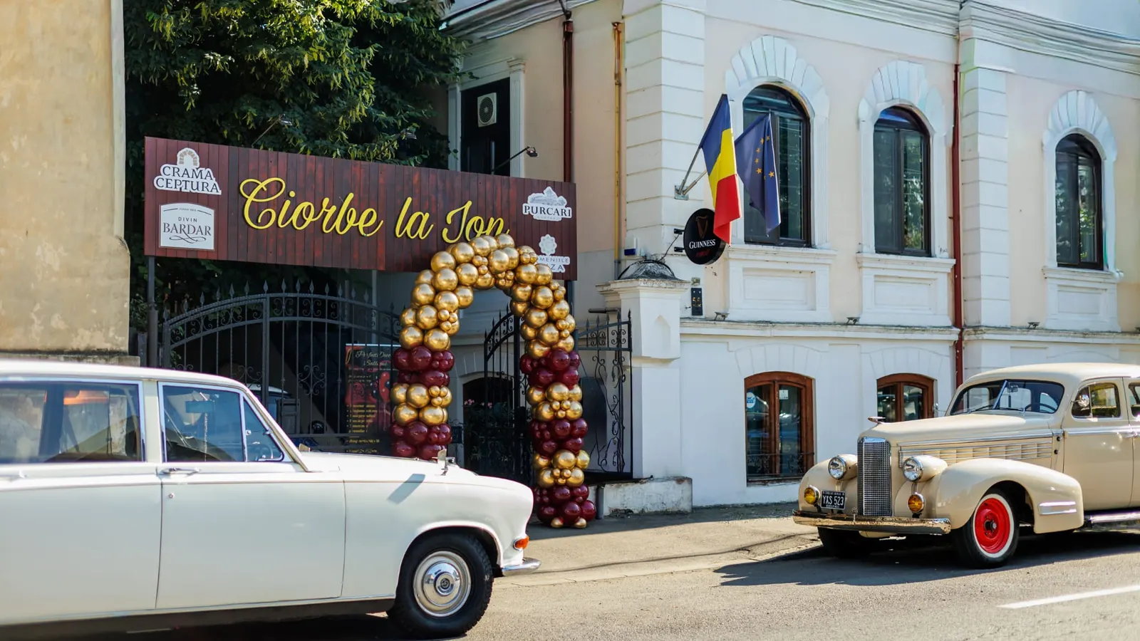 The entrance to Ciorbe La Ion, with the wooden sign and an arch of gold and burgundy balloons at the gate, between two vintage cars parked at the kerb