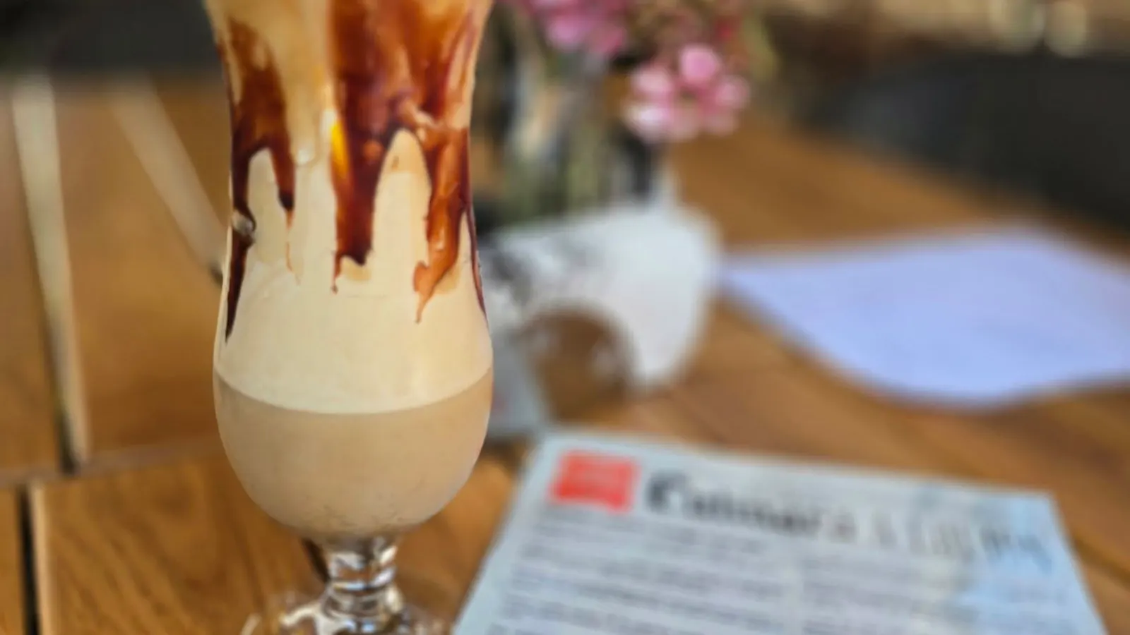 A tall glass of coffee with cream and chocolate syrup, on a wooden table